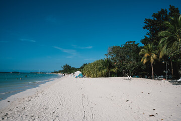 Tropical beach island paradise landscape in Nigril, Jamaica, with bright blue sky, white sand beach, palm trees, and parasols