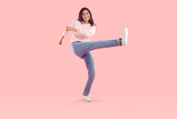 Funny carefree young woman having fun and fooling around in studio on pink background. Full length Caucasian brunette woman in stylish casual sweatshirt and jeans smiling while taking huge step.