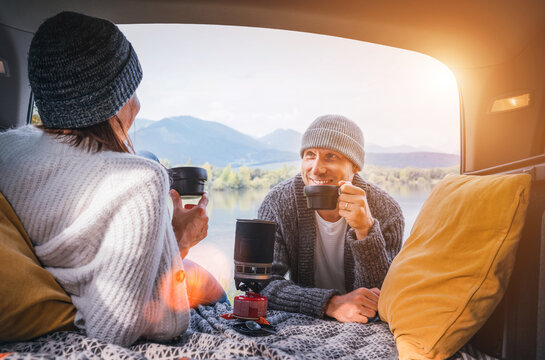 Car trunk view of chatting couple dressed warm knitted clothes enjoying gas stove prepared coffee and mountain lake view. Cozy early autumn couple auto traveling concept image.