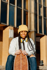 portrait looking at camera of young and latin girl with vitiligo, braids and yellow wool cap, sitting and holding a skateboard in her hands.