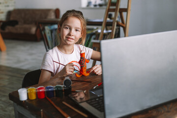 The beautiful cute girl creates crafts at home from the video lesson. A cheerful child is engaged in a hobby on a laptop. Elementary school student doing homework at homeschooling via video link.