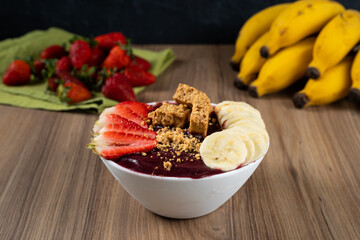 White Bowl of Brazilian Frozen Açai Berry With Paçoca, Strawberry and Banana. on a wooden desk and fruits on background