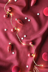 Viva Magenta color of the year 2023. Dark red Christmas background. Monochromatic flat lay with Xmas baubles, candy canes, and garland of electric festive lights on red silk textile background.