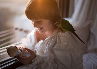 Cute Boy In White Pajamas Sitting With A Parrot On His Head Near An Open Window Against The Backdrop Of Sunset Lighting

