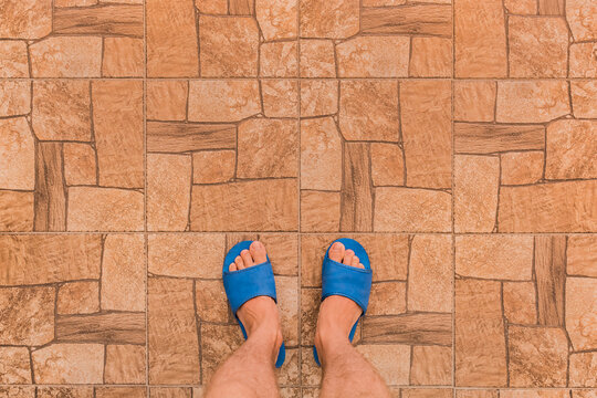 Male feet in blue house slippers stand on brown tile floor with abstract stone pattern texture background, top view