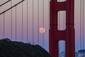 Full moon shown through the cables of the Golden Gate Bridge with purple, pink and blue sky. 