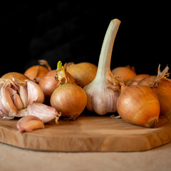 Onions and garlic lie on the table on a wooden board and a canvas napkin against black background.