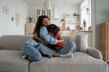 Teen girl hugging cuddling happy smiling mother, daughter showing love to mom, sitting together on couch at home. Loving parent supporting child through teenage years. Mother-daughter relationships