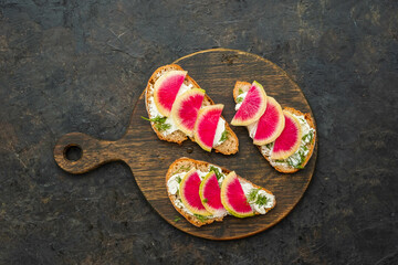 Light breakfast or snack, open sandwiches on whole grain bread with cream cheese, dill and watermelon radish slices on a wooden board against a dark concrete background. Sandwich recipes.