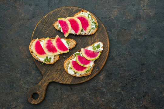 Light breakfast or snack, open sandwiches on whole grain bread with cream cheese, dill and watermelon radish slices on a wooden board against a dark concrete background. Sandwich recipes.