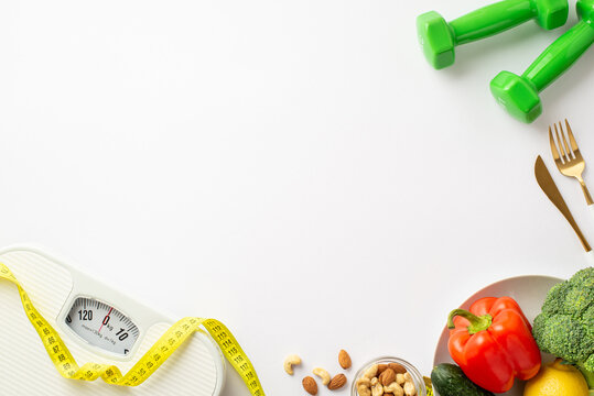 Weight loss concept. Top view photo of scales dumbbells tape measure plate with vegetables bell pepper cauliflower cucumber nuts and cutlery on isolated white background with copyspace