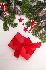 Green Christmas tree branches with pinecones decorated with red stars and Christmas candy canes, red gift centered on a white wooden background top view