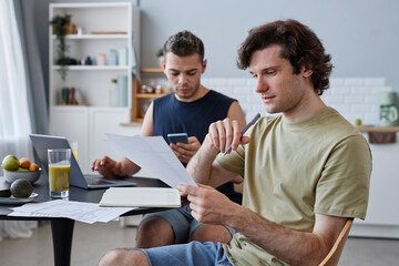 Side view portrait of young gay couple living together with focus on man studying at kitchen table in morning