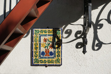 Outdoor wall of old Spanish style white stucco house, with stairway, decorative floral tile and dark, curving wrought-iron work shadows.