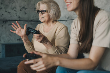 Happy daughter learning her senior mother to play video game on TV in the living room at home. Relaxing together on couch indoors. Different generations hobby