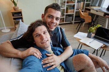 Portrait of young gay couple taking selfie photo embracing in new home, camera POV