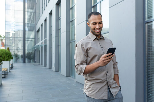Latin american businessman in casual shirt walking near office building from outside, man smiling and using phone, freelancer typing message and browsing internet pages.