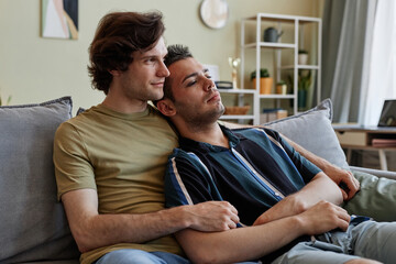 Portrait of loving gay couple relaxing on couch together and watching TV in cozy home