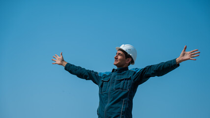 Fototapeta na wymiar A caucasian man in work clothes and a construction helmet stands against the background of a blue sky with his arms outstretched to the sides.