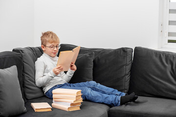 Obraz premium Child reads book in eyeglasses while sitting on sofa in room. Schoolboy studies homework.