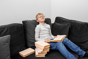 Child reads book in eyeglasses while sitting on sofa in room. Schoolboy studies homework.