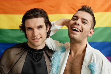Portrait of two gay young men posing with LGBTQ pride flag in background and laughing carefree