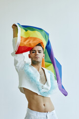 Vertical portrait of young gay man waving pride flag and wearing crop top against white