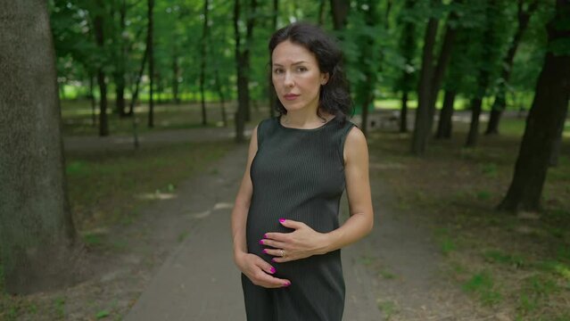 Portrait of depressed pregnant woman in black dress looking at camera with sad facial expression standing on park alley. Frustrated Caucasian lady posing in slow motion on spring summer day outdoors