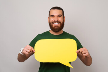 Young smiling man is holding a yellow bubble speech in front of his chest. Studio shot over grey...