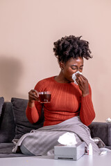 Woman sitting on a sofa, feeling sick. African American girl having flu, blowing her nose, and...