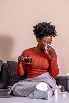 Woman sitting on a sofa, feeling sick. African American girl having flu, blowing her nose, and holding a cup of tea.