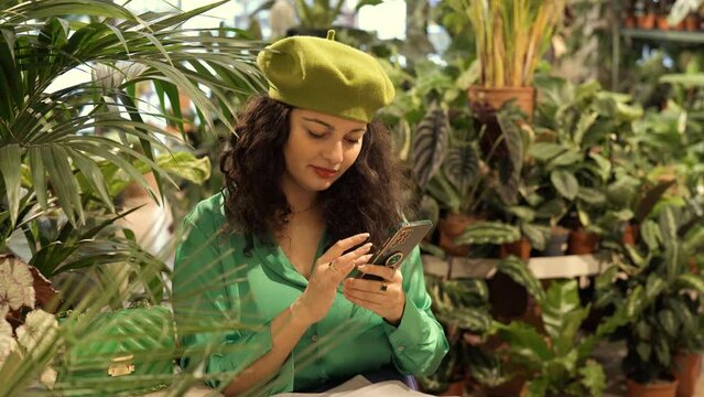 Stylish hipster woman using mobile phone in a cage with many green house plants. Hygge interior design. Biophilic lifestyle
