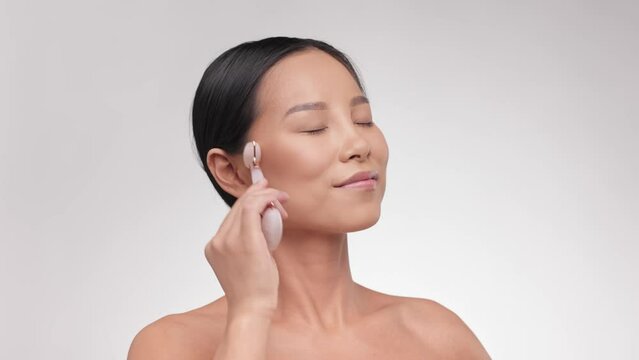 Face massage. Portrait Smiling young asian woman using jade facial roller for skin care, beauty treatment on white isolate background. Young Girl using natural massager. Skin care concept.