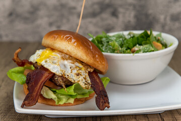 Huge combination breakfast meal burger stacked high with bacon, eggs, tomato, and served with a bowl of caesar salad