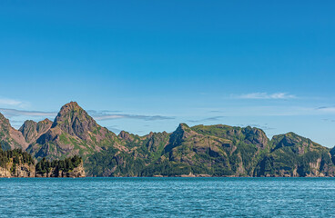 Naklejka premium Resurrection Bay, Alaska, USA - July 22, 2011: Partly green by foliage, a brown rocky mountain range creates a belt between blue water and a blue cloudscape