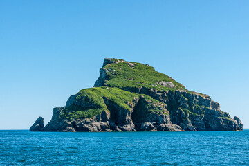 Resurrection Bay, Alaska, USA - July 22, 2011: Closeup of Hive island shows green slanted surface ending in gray steep cliffs into deep blue ocean water under light blue sky