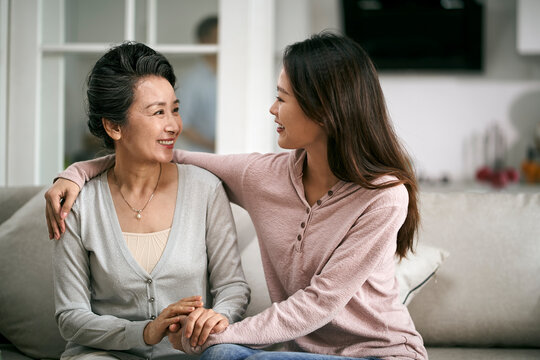 asian adult daughter and senior mother sitting on couch at home chatting talking conversing