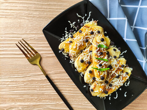 Grilled banana topped with grated cheese and chocolate chips. Served on black plate on wood textured background. Selective focus image.
