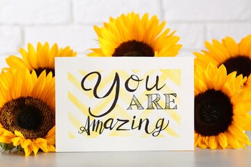 Card with life-affirming phrase You Are Amazing and sunflowers on light table against white brick...