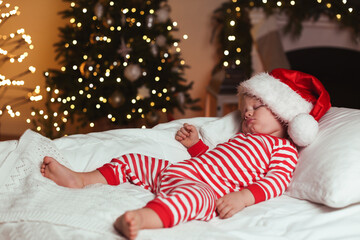 Fototapeta na wymiar Baby in Christmas pajamas and Santa hat sleeping on bed indoors. Space for text