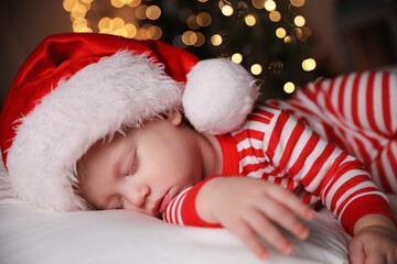 Baby in Christmas pajamas and Santa hat sleeping on bed indoors