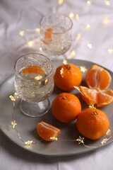 Delicious ripe tangerines, glasses of sparkling wine and fairy lights on white bedsheet
