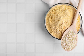 Sesame flour in glass bowl with wooden spoon of seeds on white tiled table, top view. Space for text