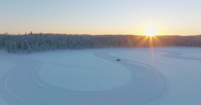 Driver drifting corners on Lapland ice lake race track as sunrise rises from woodland aerial view