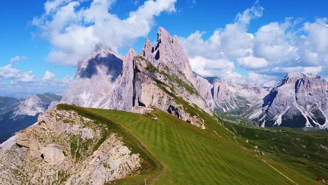 Pull back aerial view from of the peaks of Seceda with green pastures, and hiking trails in the foreground, and mountain peaks in the background in the Italian Dolomites in South Tyrol, Italy