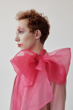 Side view portrait of young man with creative make-up wearing big pink bow standing against white wall background