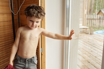 Little kid standing shirtless resting against window horizontal medium portrait