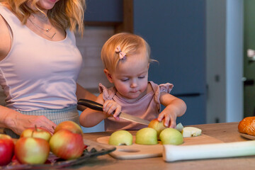 Fototapeta premium A little girl cooks in the kitchen with her mother. A child in a pink sweater cuts apples. Close-up. Love and tenderness.