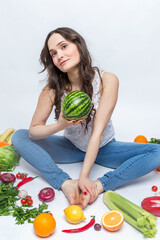 Fototapeta premium A young woman sits and holds a watermelon in her hands. Smiling brunette in jeans. Pile of vegetables and fruits on the floor. Healthy food and vegetarianism. White background. Vertical.