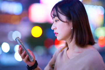 夜の繁華街でスマートフォンを見る若い女性